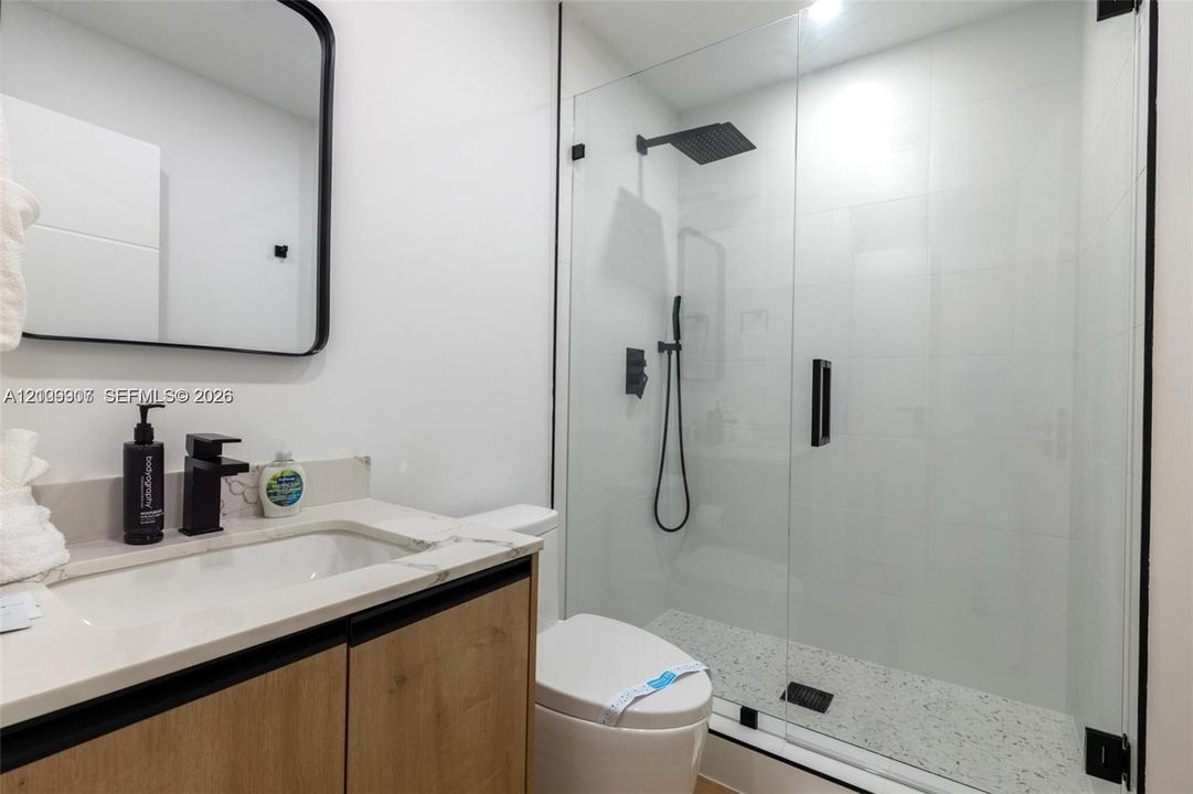 Bathroom featuring a wood-finish vanity with a stone-surface countertop, a frameless glass shower enclosure, matte black fixtures, a rectangular vanity mirror, and a speckled-finish shower floor