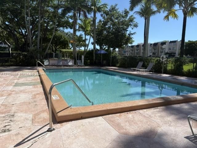 Rectangular swimming pool with metal handrails, surrounded by stone-paver decking, mature palm trees, and perimeter landscaping