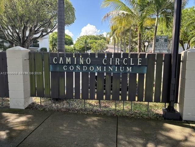 Community entrance featuring painted wood fencing, masonry pillars, and mature palm tree landscaping