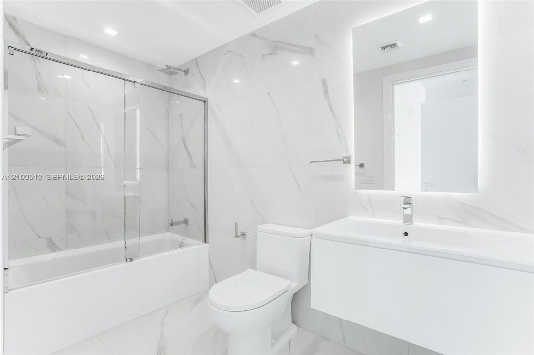 Bathroom featuring large-format porcelain tile, wall-mounted floating vanity, glass tub enclosure, and backlit vanity mirror