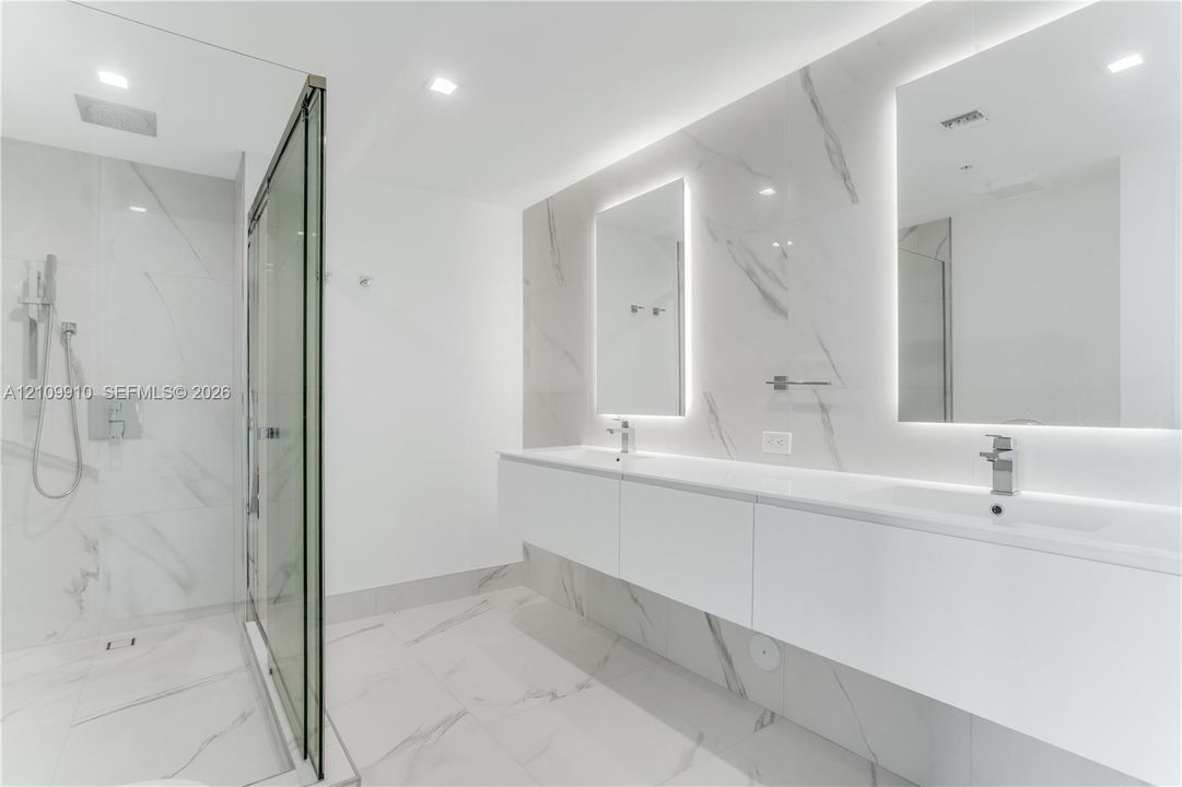 Floating double vanity with dual rectangular sinks, floor-to-ceiling stone-finish tile wall, glass-enclosed walk-in shower with rainfall fixture, and recessed LED-backlit mirrors