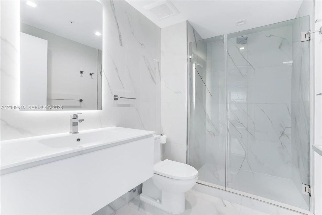 Bathroom featuring a floating vanity with a smooth surface, wall-to-wall mirror with integrated lighting, large-format stone-patterned wall tile, and a glass-enclosed shower
