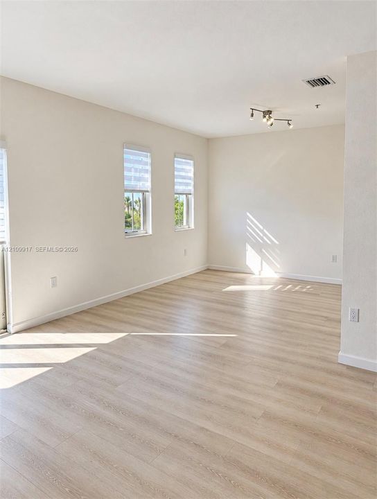 Neutral interior space featuring wood-finish flooring, crisp white walls, dual windows with roller shades, and a multi-directional track lighting fixture