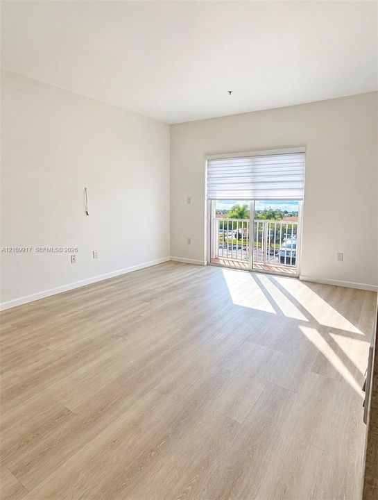 Light-filled room featuring wood-finish flooring, neutral wall paint, and sliding glass doors with an integrated shade opening to an exterior balcony