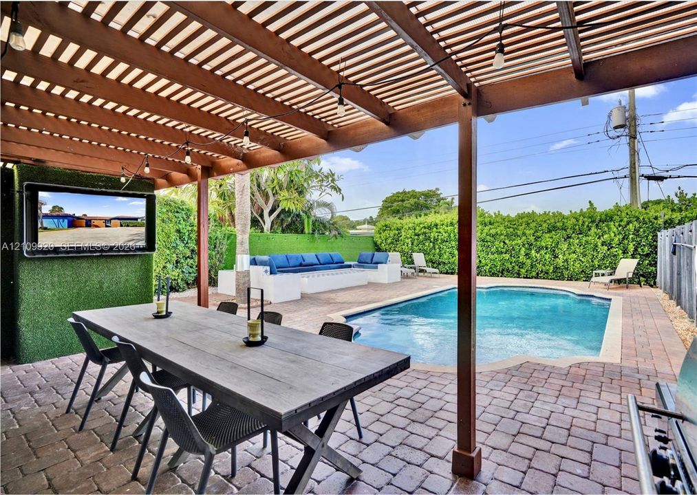 Residence 1747 (Left Side) – Covered outdoor patio featuring a wooden pergola, new flat roof, brick paver flooring, heated pool, built-in wall-mount TV.