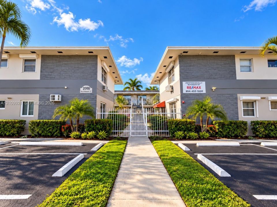 Multi-unit exterior featuring gray stone-veneer accents, paved parking area, gated pedestrian entrance, and manicured landscaping
