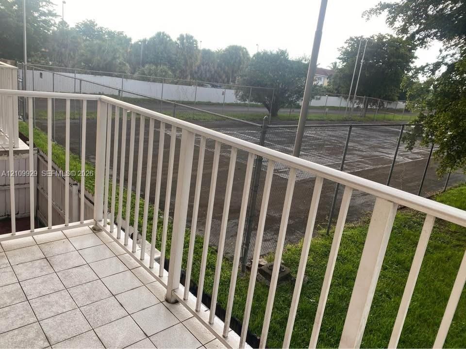 Exterior balcony featuring white metal railings and tiled flooring, overlooking an enclosed tennis court