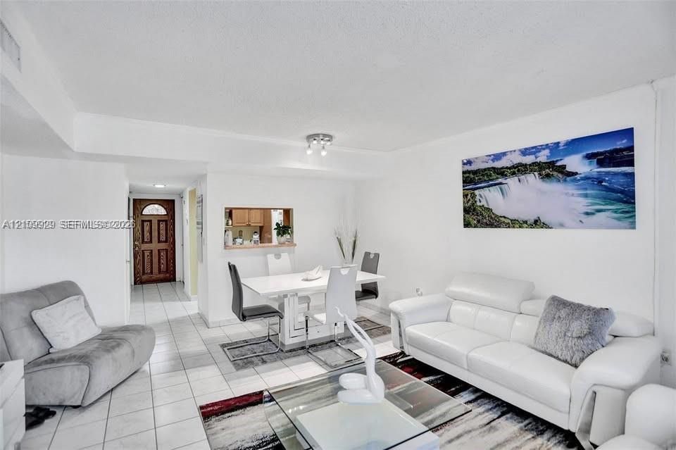 Open-concept living space with white ceramic tile flooring, built-in kitchen pass-through window, and multi-light flush-mount ceiling fixture, featuring a decorative wood-panel entry door with inset glass detailing