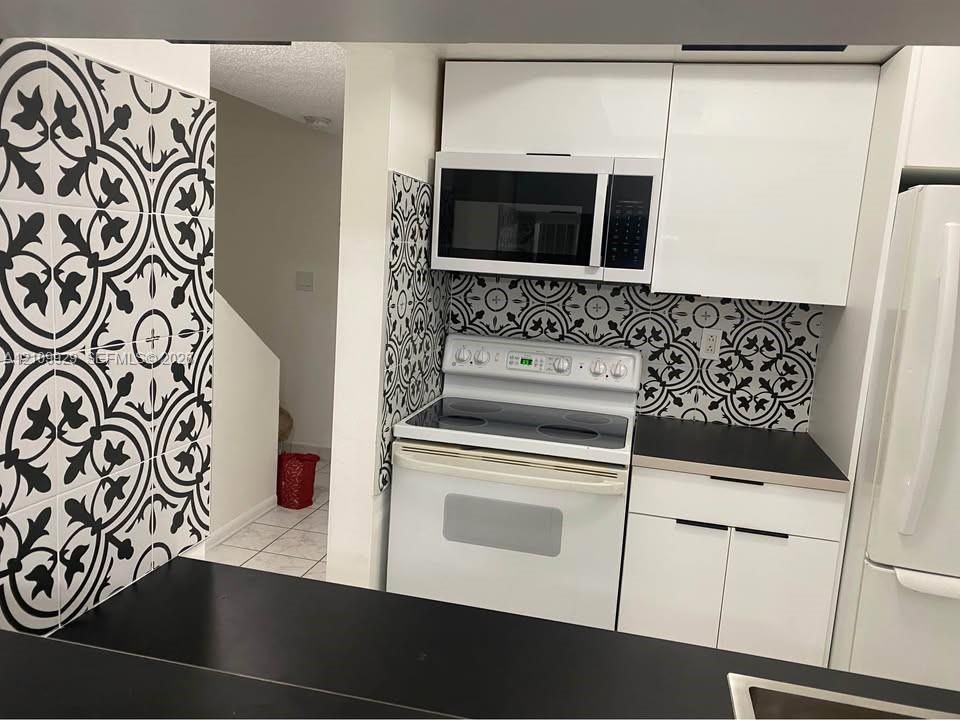 Kitchen featuring white flat-panel cabinetry, a geometric patterned tile backsplash, a black countertop, and an integrated white electric range and microwave
