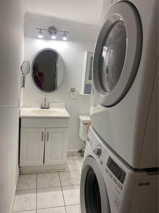 Vanity featuring a white cabinet, integrated countertop with sink, and a multi-bulb light fixture above an oval mirror, complemented by stone-finish tile flooring, a wall-mounted storage cabinet, and a stacked laundry unit