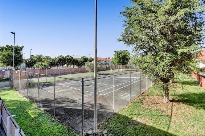 Fenced tennis court featuring a hard surface, mesh perimeter enclosures, integrated lighting poles, and surrounding lawn areas with mature trees