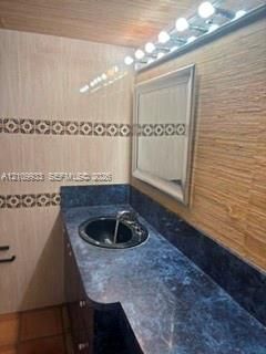 Vanity featuring a dark stone-finish countertop, integrated basin with metal hardware, tiled walls with decorative inlay, textured wood-finish wall paneling, and a multi-bulb vanity light fixture