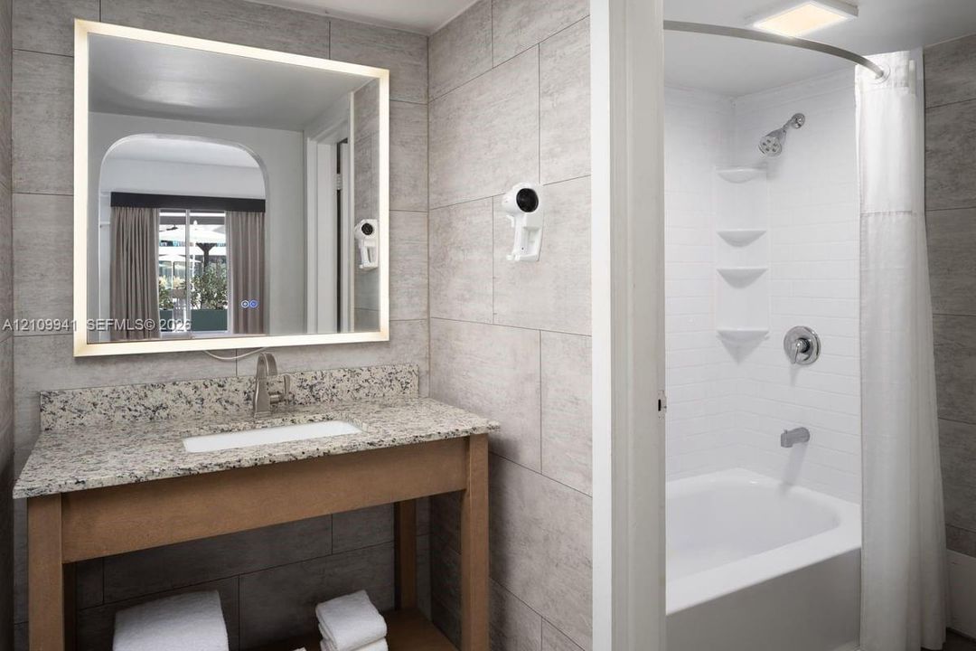 Bathroom featuring a stone-top vanity with an integrated rectangular sink, wall-mounted illuminated mirror, large-format tile wall surfacing, and a shower-tub combination with a curved curtain rod