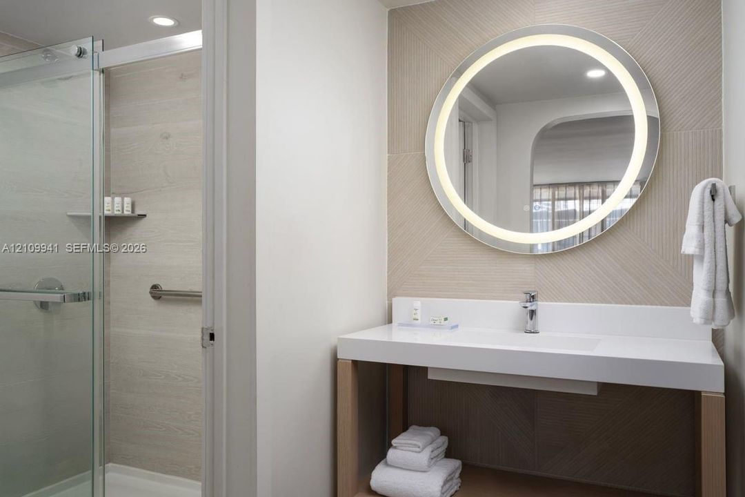 Modern vanity with a seamless integrated sink, wall-mounted circular LED mirror, glass-enclosed shower, and neutral patterned wall tile