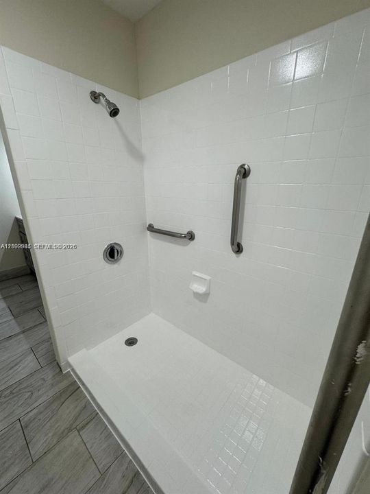 Walk-in shower featuring white tiled surround, built-in soap dish, metallic shower head, and stainless steel grab bars, complemented by wood-finish floor tiling
