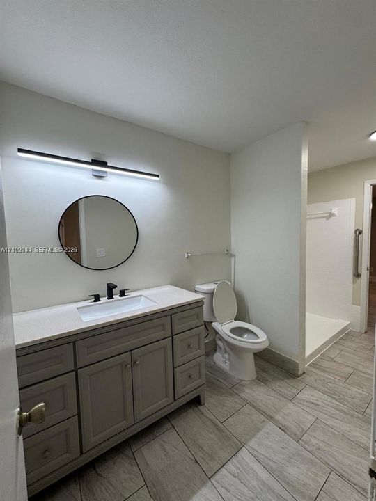 Bathroom featuring a grey shaker-style vanity with a solid surface countertop, rectangular undermount sink, matte black widespread faucet, and circular wall-mounted mirror with integrated linear lighting, complemented by tile flooring