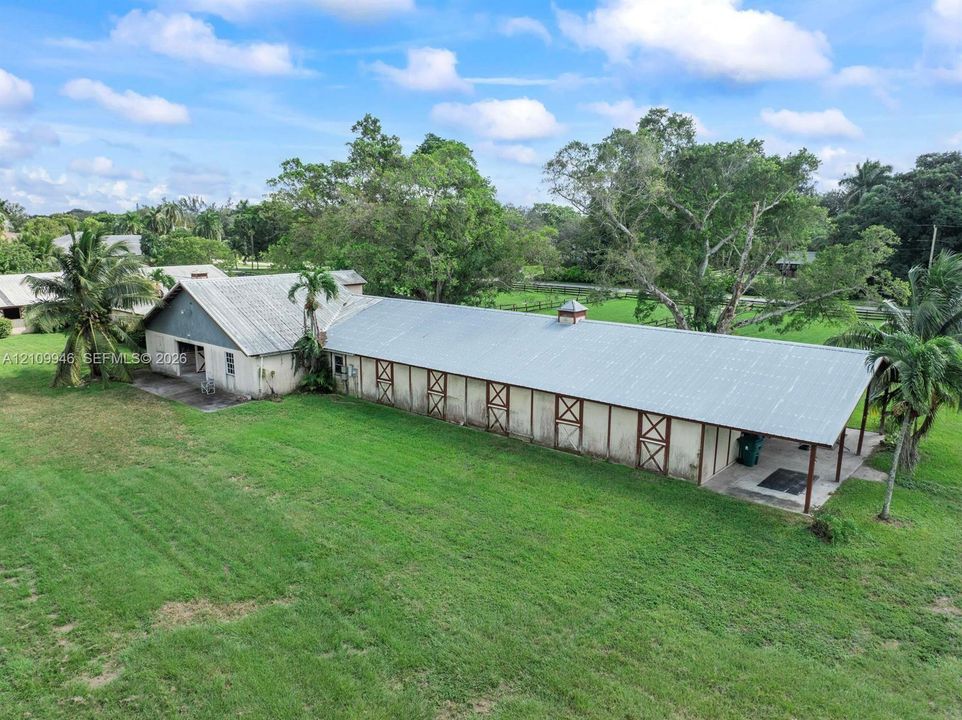 Extensive barn structure featuring a metal roof, multiple exterior stall doors, a covered porch area, and manicured surrounding acreage