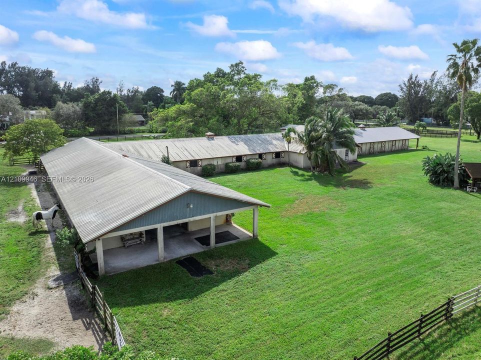 L-shaped stable facility featuring metal roofing, multiple stalls, integrated covered overhangs, and perimeter wood-rail fencing situated on expansive acreage