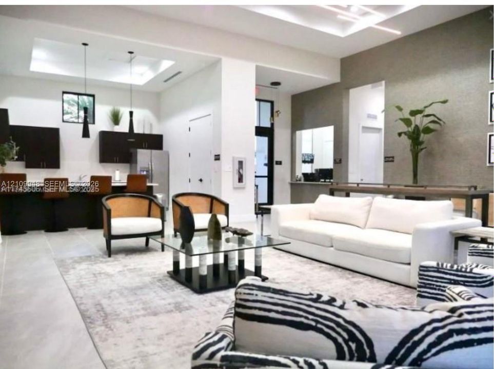 Open-concept space featuring recessed tray ceilings with integrated linear lighting, large-format tile flooring, kitchen with dark cabinetry and peninsula seating, and a decorative textured accent wall