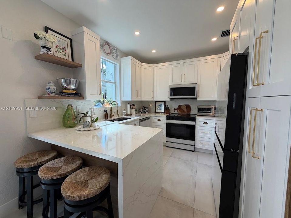 Kitchen featuring white shaker-style cabinetry with gold-tone hardware, waterfall-edge stone-composite countertops, tile flooring, built-in stainless steel appliances, and wood-finish floating shelving