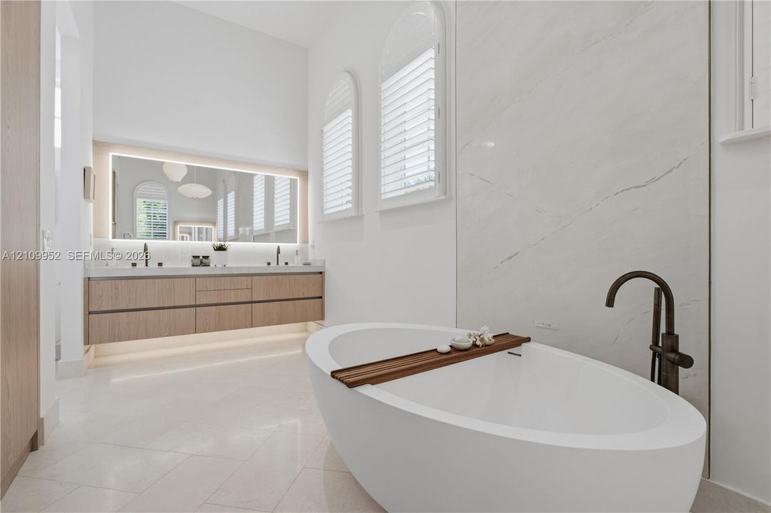 Freestanding oval bathtub with matte black floor-mounted faucet, wall-to-wall stone slab surround, floating wood-finish vanity with dual sinks, backlit mirror, and arched windows with plantation shutters