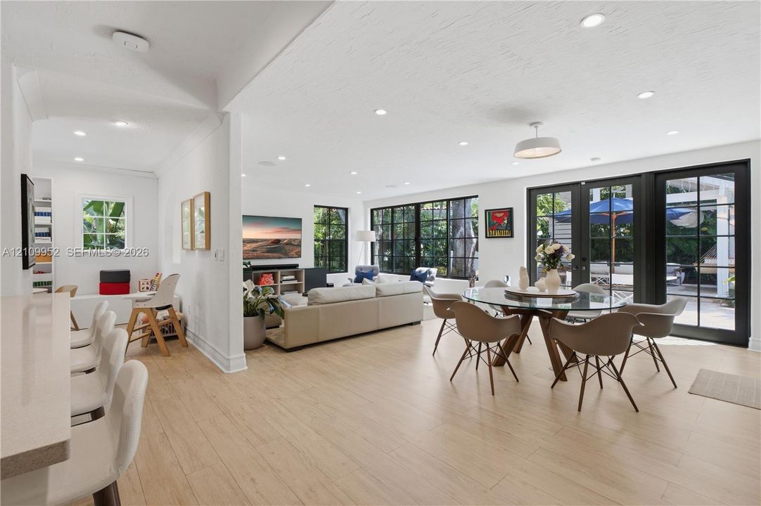 Open-concept interior featuring light wood-finish flooring, recessed lighting, expansive multi-pane black-trimmed windows, and double black-trimmed French doors leading to the exterior