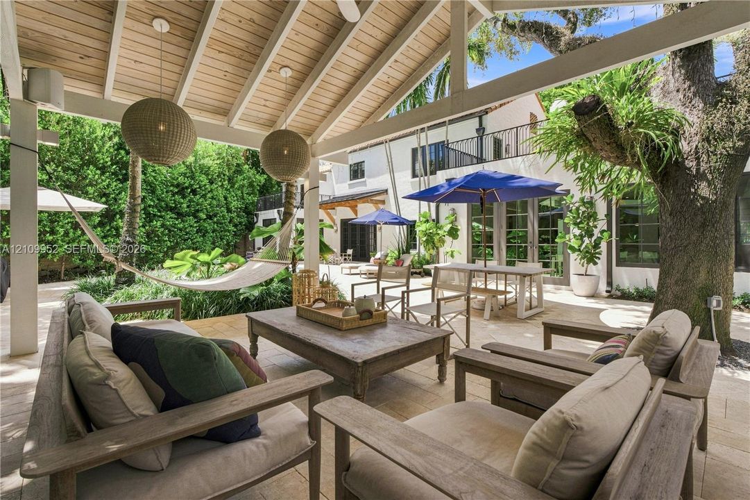 Covered patio featuring a beamed wood ceiling, stone tile flooring, pendant lighting, and direct access to a hardscaped outdoor area with mature trees