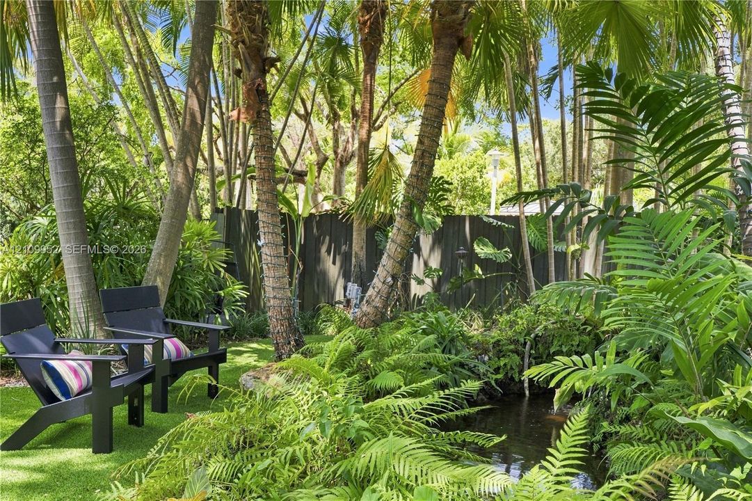 Landscaped garden featuring mature palm trees, a pond, lush tropical foliage, and a dark wood-finish privacy fence