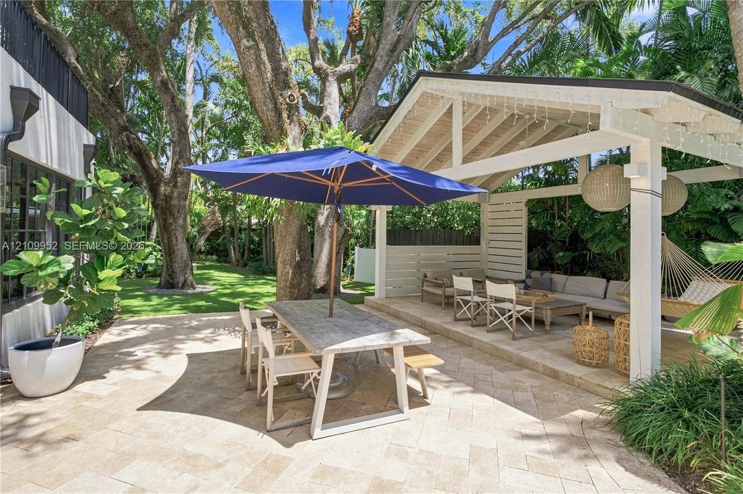 Stone-paver patio area featuring a white timber-frame pergola, mature trees, manicured lawn, and privacy fencing