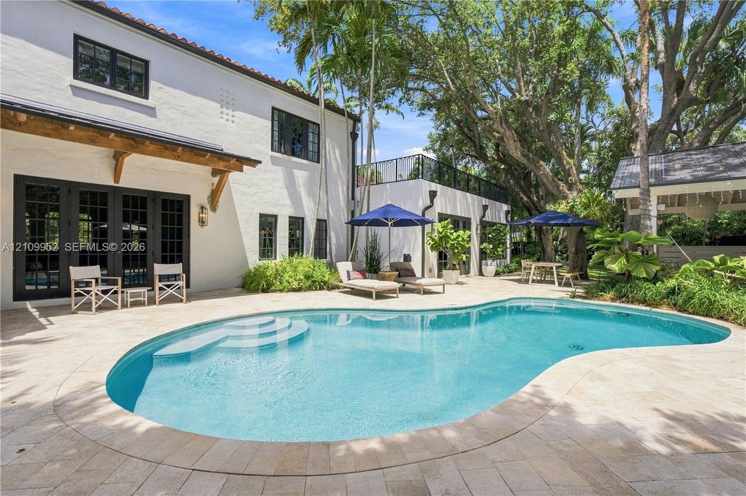 Stucco exterior featuring a curved pool with stone paver decking, a second-story terrace with a metal railing, wood-beam architectural accents, and mature shade trees