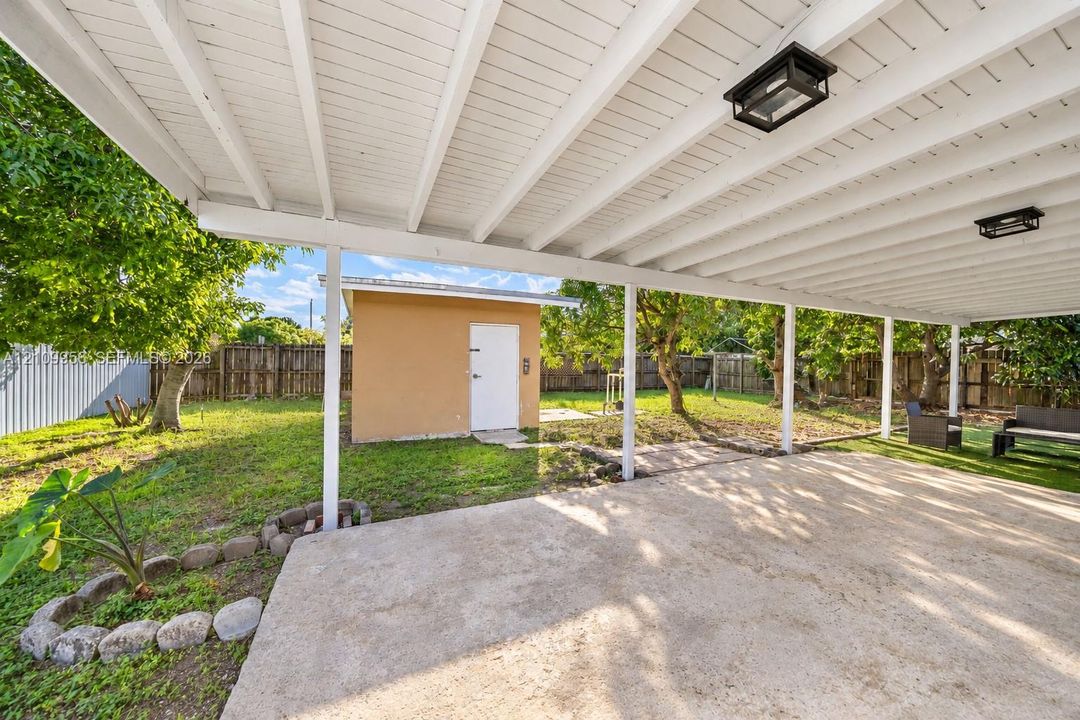 Covered concrete patio with overhead lighting, detached storage building, and fenced backyard with mature trees