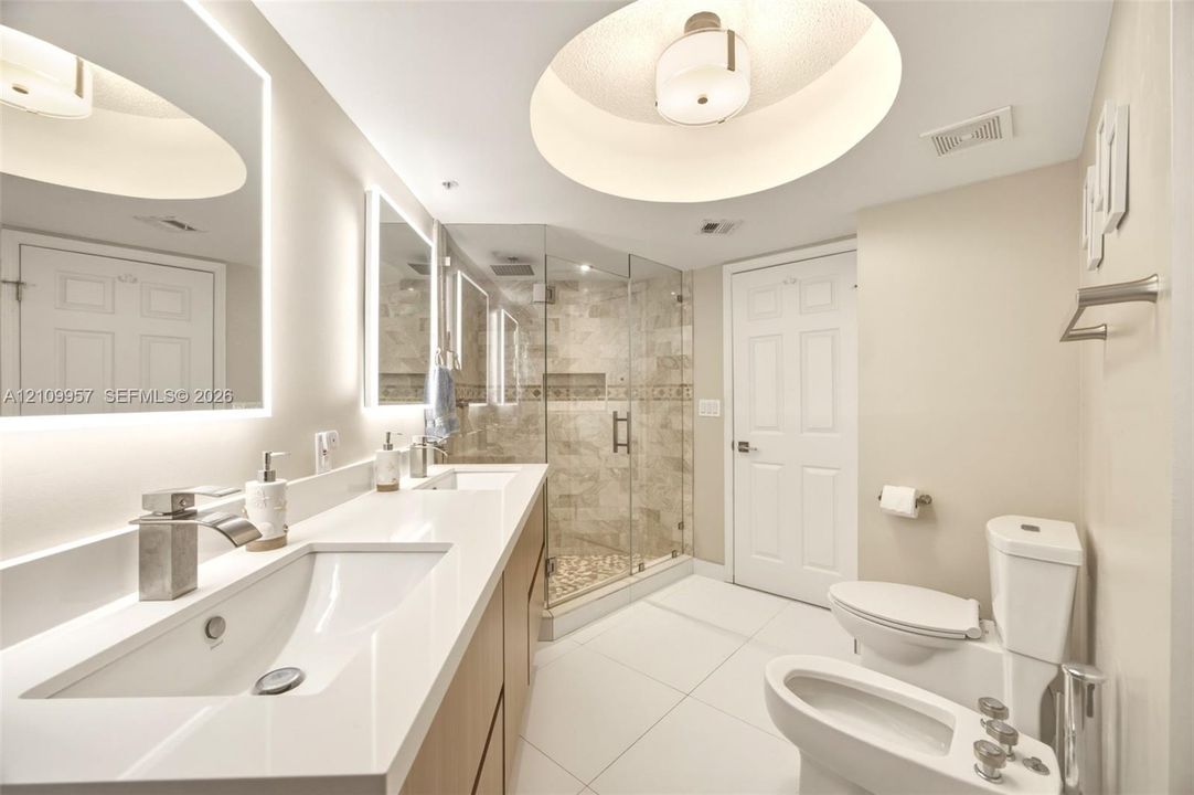 Newly updated primary bathroom with dual-integrated sinks and quartz countertop, backlit mirrors, freestanding soaking tub, wood-finish tile accent wall, and bidet