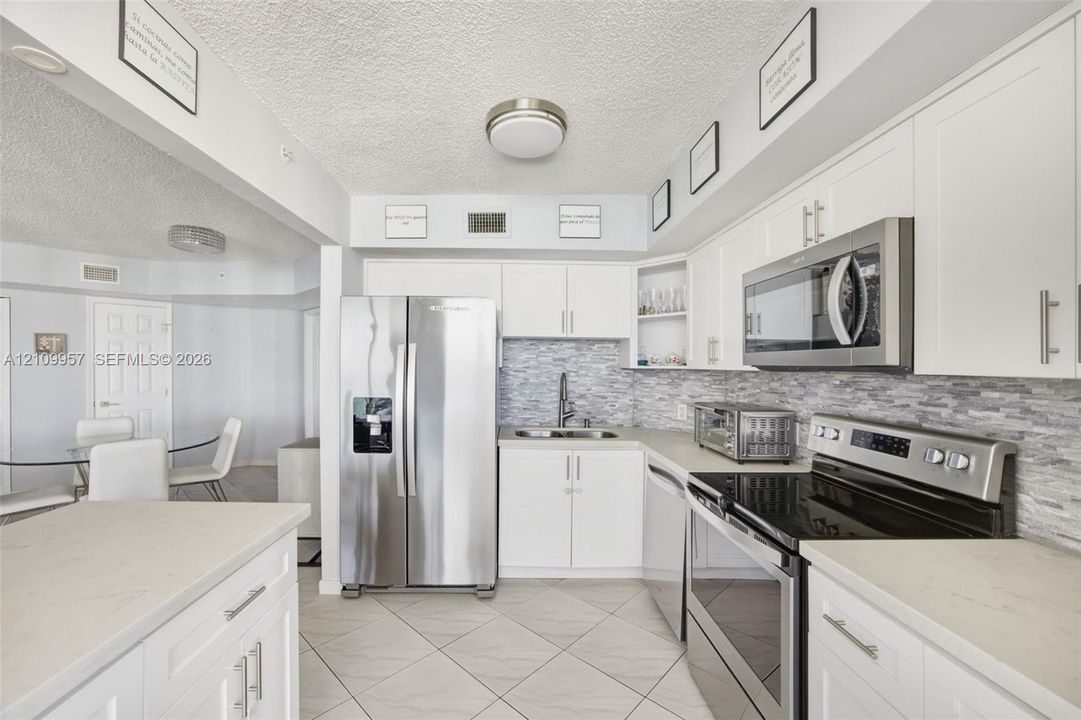 Updated kitchen featuring white shaker-style cabinetry, stainless steel appliances, light-toned stone-finish countertops, a multi-tonal stacked stone backsplash, and large-format tile flooring