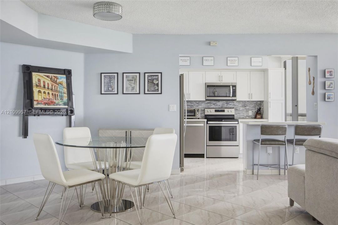 Updated kitchen featuring white cabinetry, stainless steel appliances, a stone-patterned tile backsplash, and a breakfast bar, complemented by neutral tile flooring