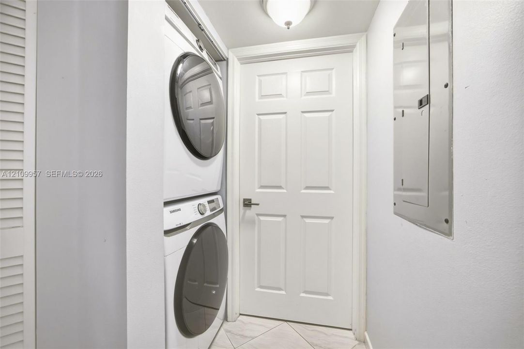 Laundry closet featuring a stacked front-load washer and dryer set, neutral-toned tile flooring, and a six-panel door with a brushed nickel lever