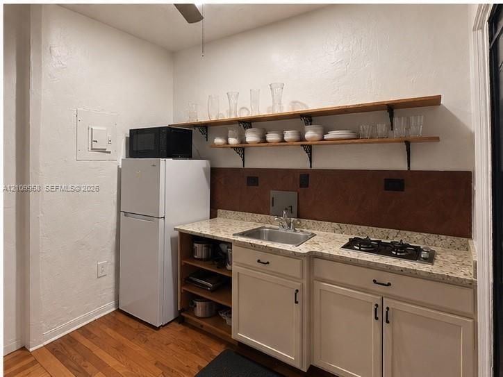 Kitchenette featuring neutral-toned cabinetry, light-colored stone-finish countertops, an integrated gas cooktop, a stainless steel sink with a high-arch faucet, and dual wooden floating shelves