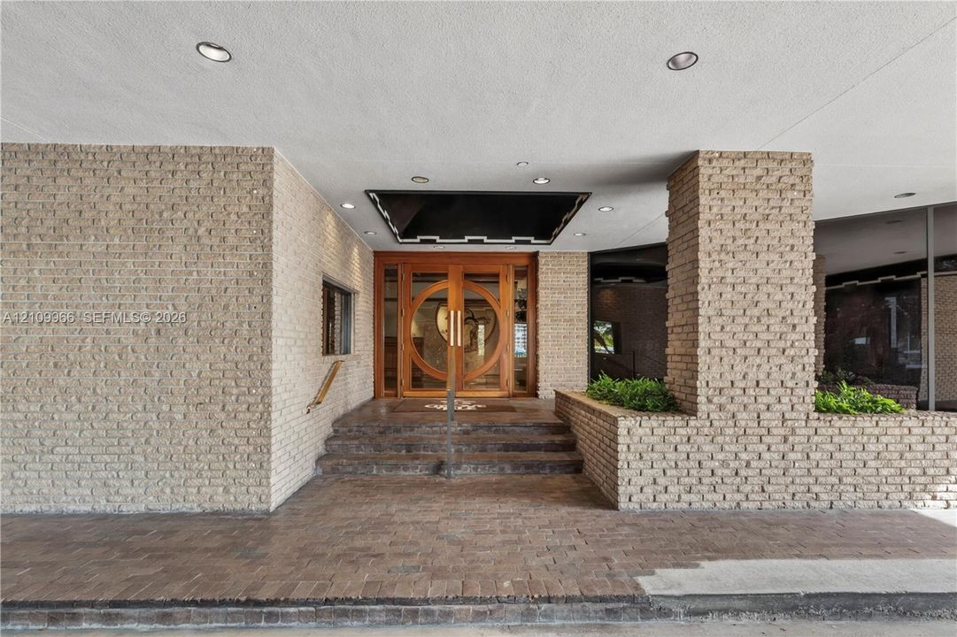 Building Exterior entrance featuring light-toned brick masonry, stone steps, dual wood-framed circular glass doors, recessed ceiling lighting, and integrated brick planters