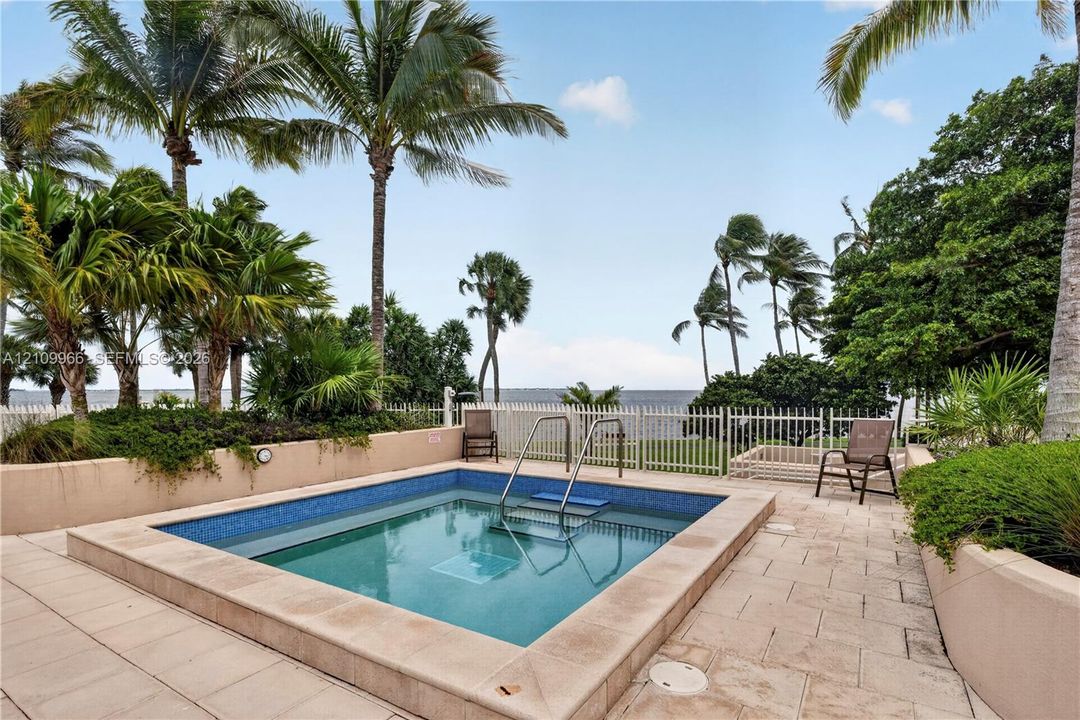 In-ground pool featuring blue tile inlay, stone-finish coping, metal handrails, and a surrounding light-toned stone deck with mature palm trees