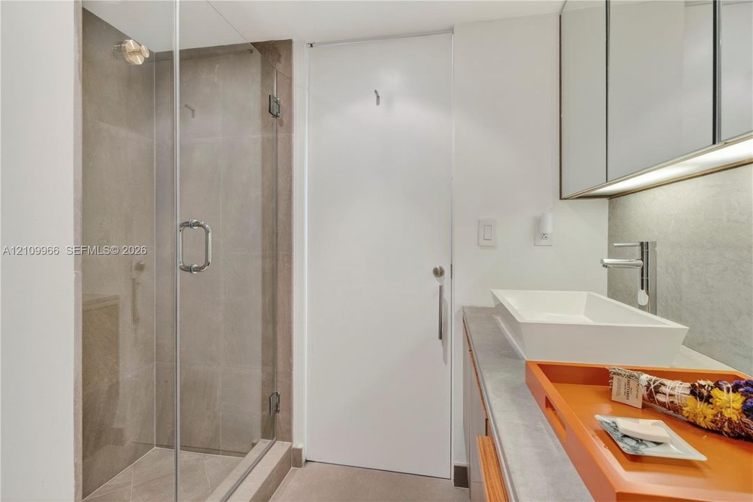 Walk-in shower with frameless glass enclosure, rectangular vessel sink, polished chrome faucet, stone-finish countertop, and wall-mounted mirrored cabinetry