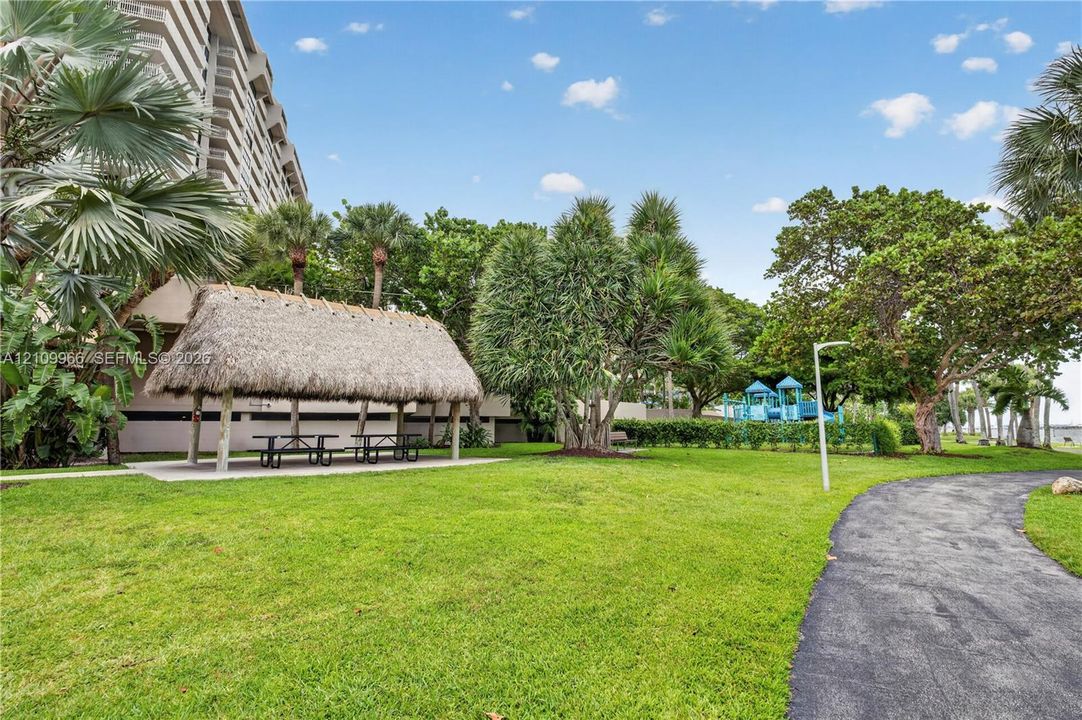 Landscaped grounds featuring a thatched-roof pavilion, paved walking paths, mature palm trees, and a play structure