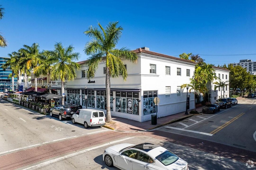 Corner commercial building featuring white stucco facade, expansive ground-level storefront windows, decorative roofline cornice, and mature palm trees