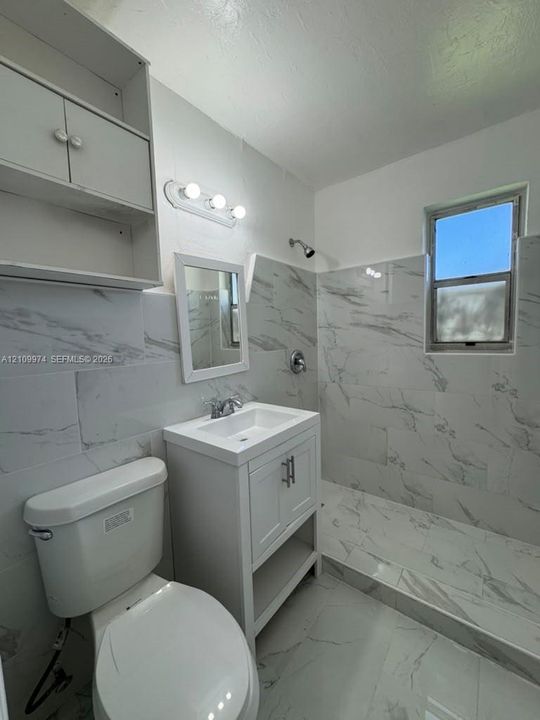 Full-height tile shower enclosure with stone-patterned surfacing, integrated vanity with lower open shelving, overhead built-in cabinetry, polished chrome fixtures, and neutral tile flooring