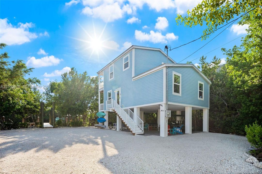 Elevated two-story residence with light blue horizontal siding, open-air ground level, exterior staircase with white railings, and gravel driveway