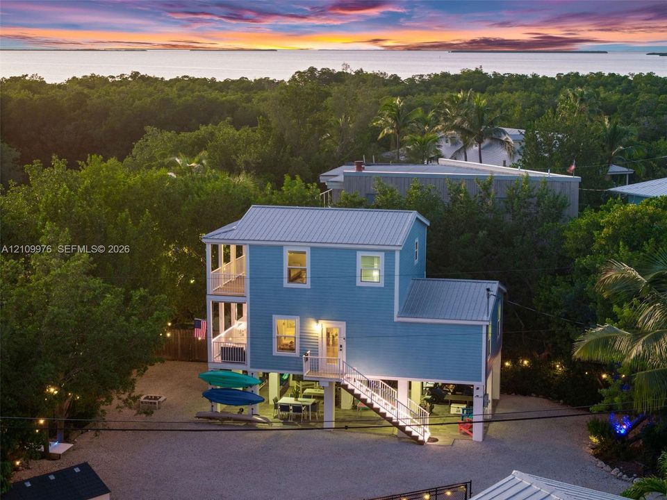 Elevated coastal residence featuring a metal roof, multi-level covered porches, and a gravel ground-level space, set against a backdrop of mature foliage and open water