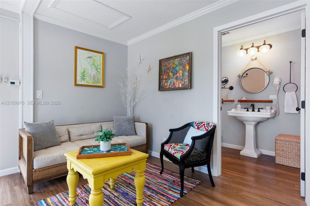 Interior space featuring wood-finish flooring and crown molding, with an opening to a bathroom equipped with a pedestal sink, three-bulb vanity lighting, and a circular mirror suspended by rope