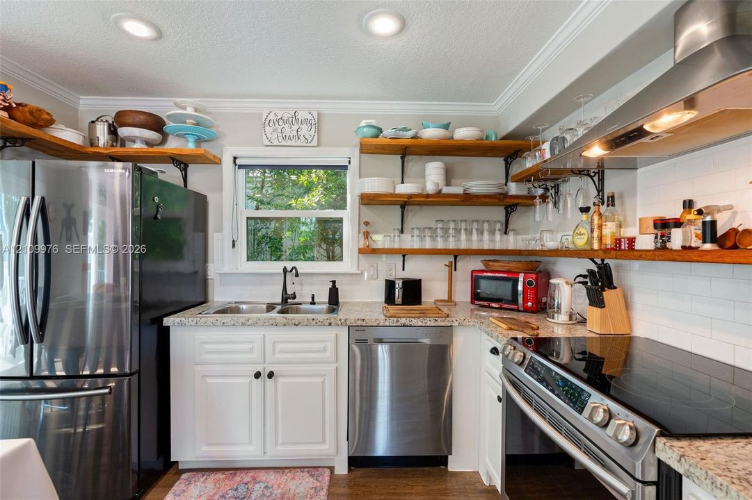 Kitchen featuring stainless steel appliances, speckled stone countertops, white cabinetry, open wood shelving, and a glass-tiled backsplash