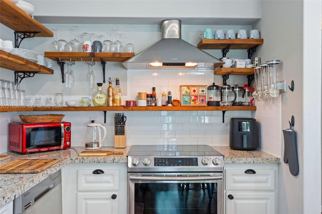 Kitchen area featuring white shaker-style cabinetry, natural stone countertops, white subway tile backsplash, stainless steel range with overhead vent hood, and open wood shelving