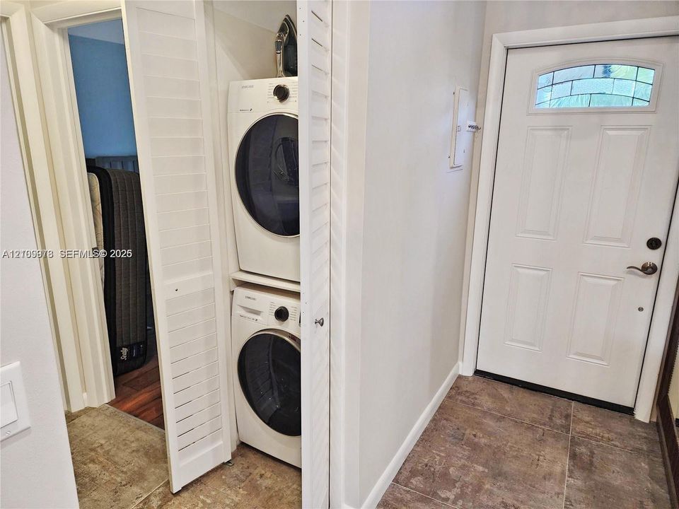 Laundry closet featuring stacked front-load washer and dryer, louvered bi-fold doors, and a exterior-facing door with an arched glass inset