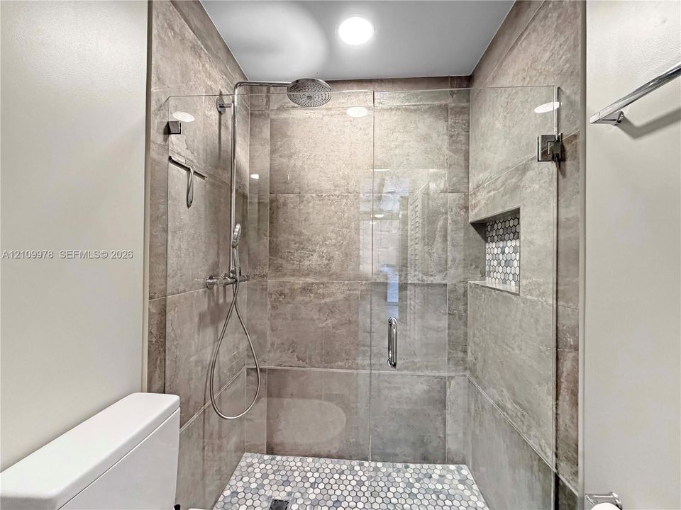 Walk-in shower featuring large-format neutral wall tile, mosaic penny tile flooring, integrated shower niche, frameless glass enclosure, and dual-head rain-style fixture