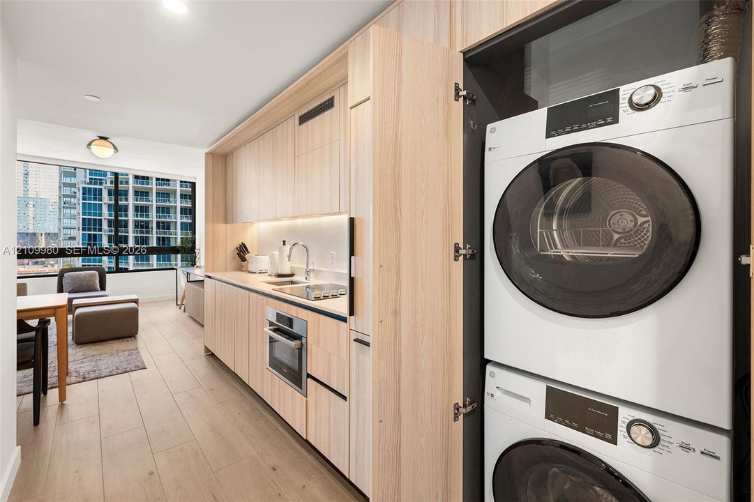 Kitchenette featuring light-toned cabinetry, built-in stacked laundry unit, integrated under-cabinet lighting, and wood-finish flooring